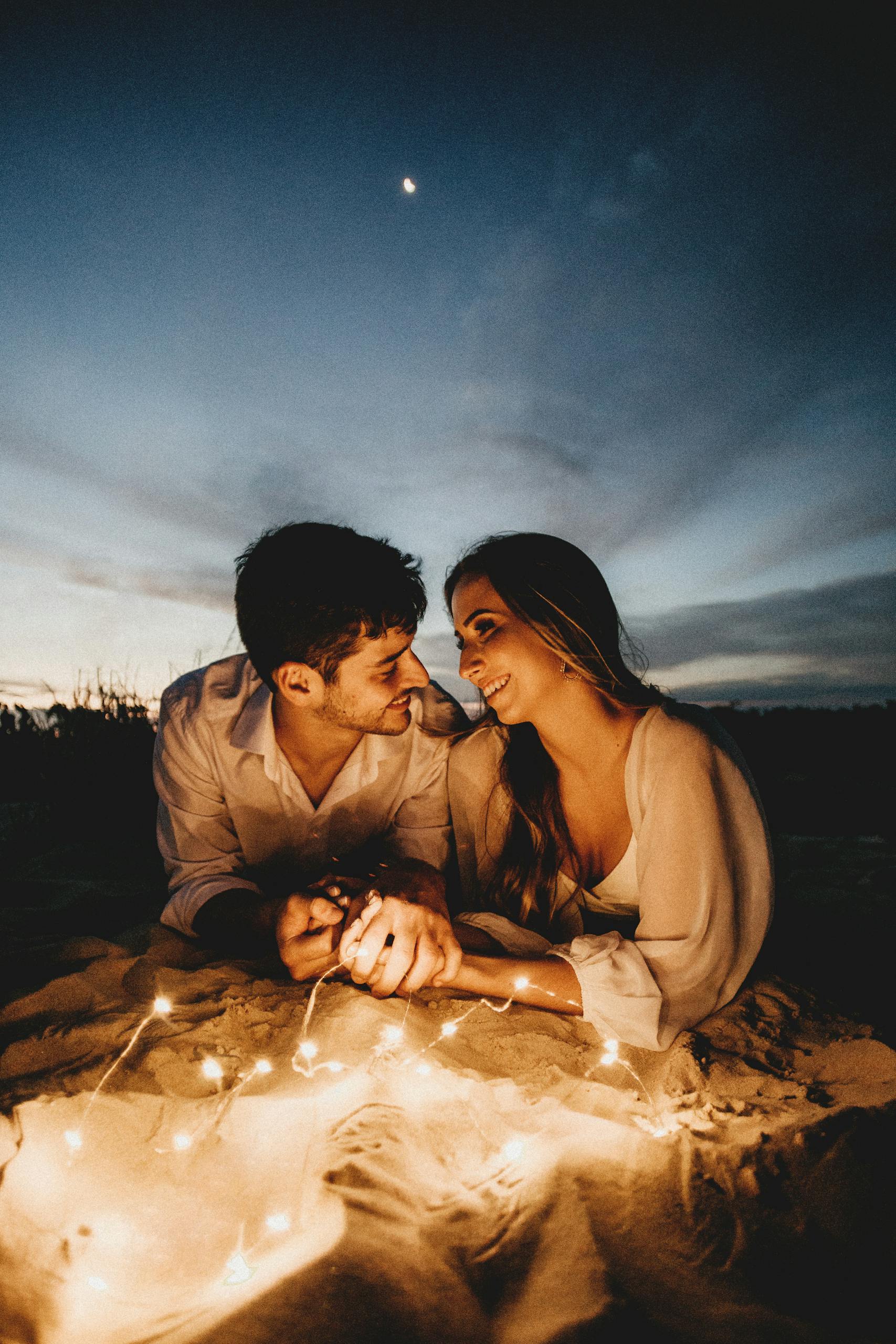 A loving couple lying on sandy ground, illuminated by fairy lights under a starry twilight sky.