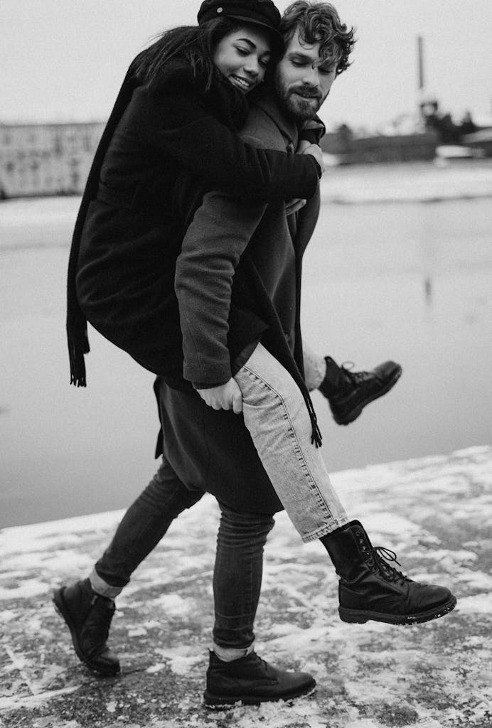 Black and white photo of a happy couple enjoying a piggyback ride by a snowy riverbank.