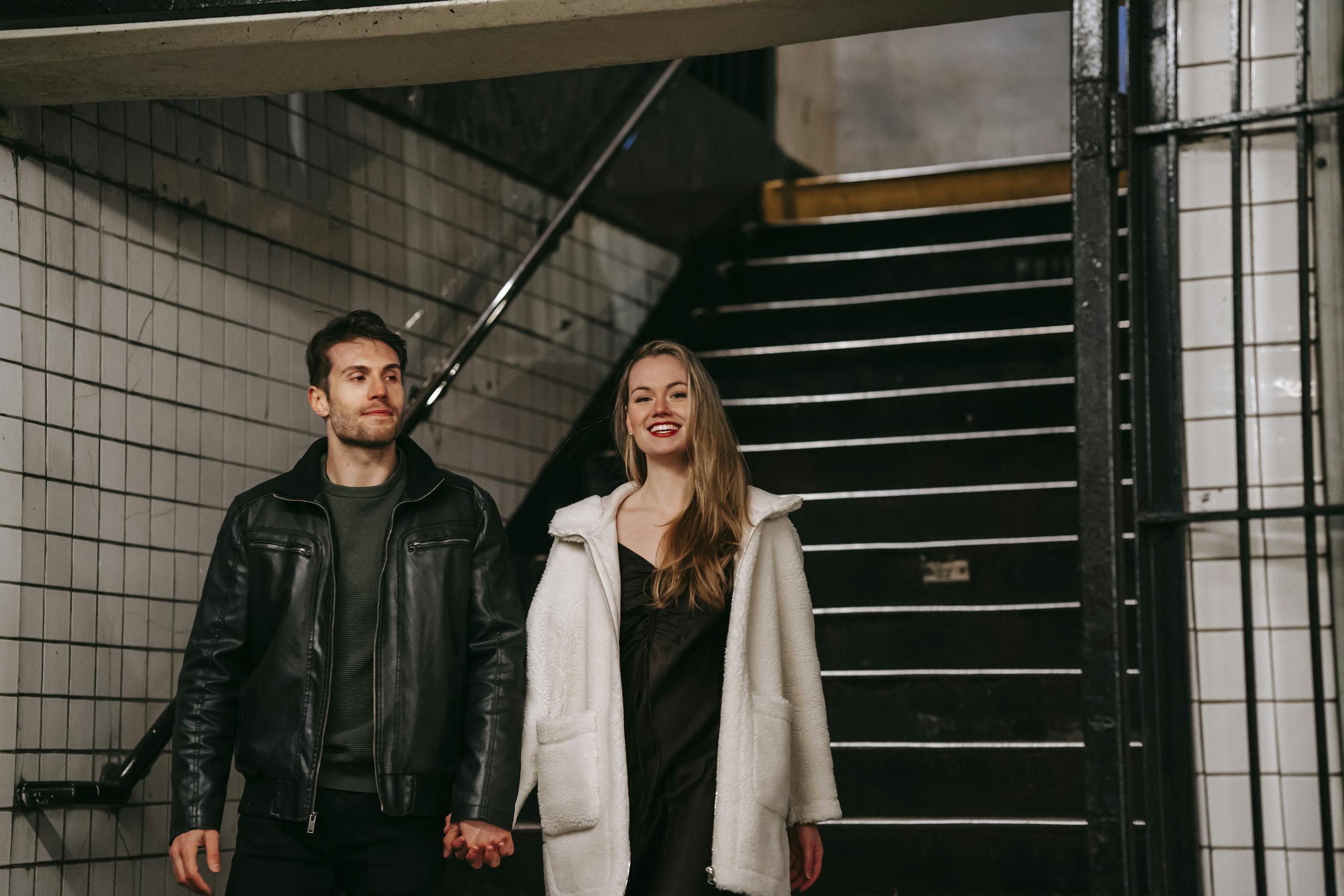 Cheerful couple in casual clothes holding hands and going downstairs in underground passage placed in city in evening time